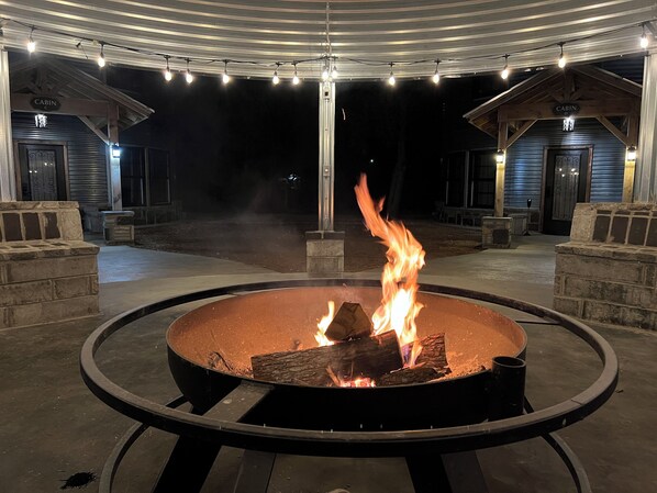 Ein Außenfeuerbereich mit einer Sitzordnung im Kreis, umgeben von Steinbänken und einem überdachten Pavillon mit Lichterketten.