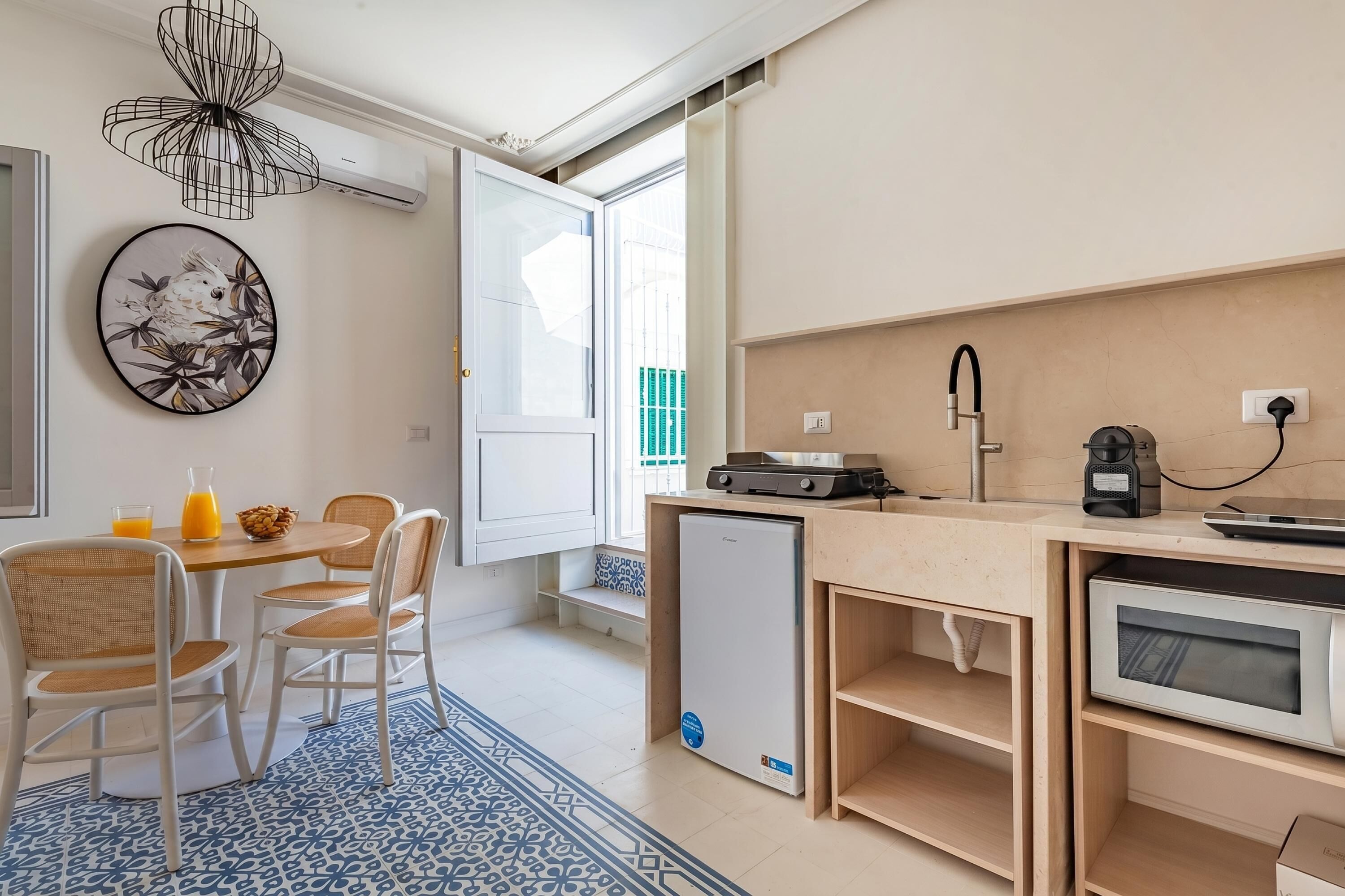 A compact kitchen with a refrigerator, microwave, and coffee maker, a dining area with chairs and a table, and a wall-mounted air conditioner.