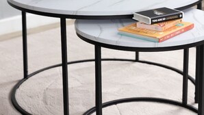 A round, white marble table with black metal legs, accompanied by a smaller table. A potted plant and a stack of books are on the main table. A television and a wooden sign are in the background.