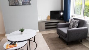 A living room with a grey armchair, a small round table with a plant, a flat-screen TV on a wooden stand, and a window with a view of greenery.
