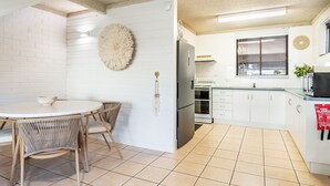 A kitchen with white brick walls, a round dining table with chairs, a refrigerator, an oven, and a window with blinds. - Pet-friendly 2 bedroom villa (Lennox Head)
