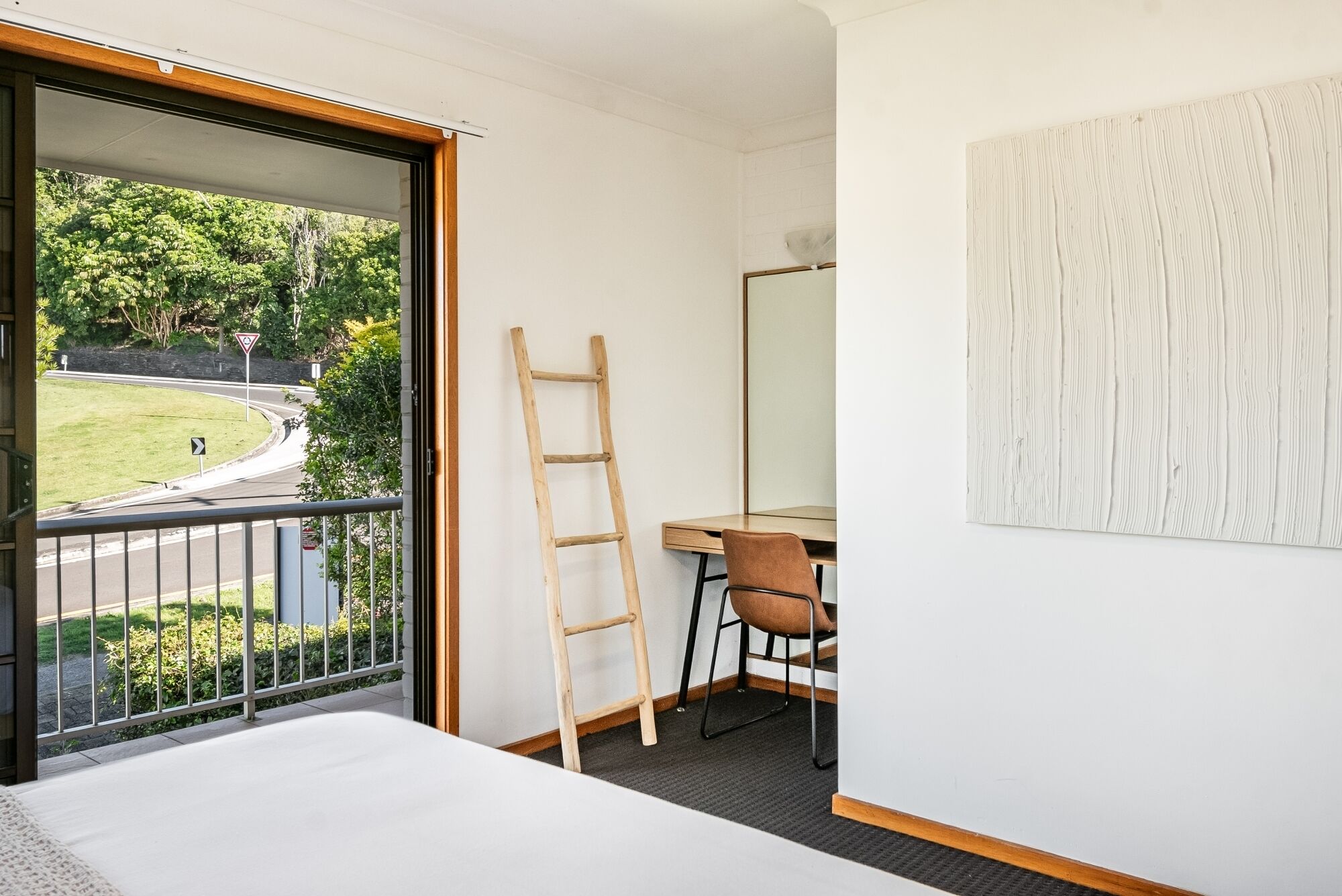 A room with a bed, a desk with a chair, a ladder, and a large window overlooking a road and greenery.