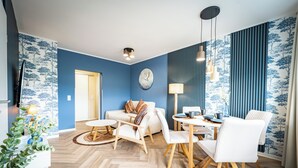 A modern living room with a blue wall, a sofa, a dining table with chairs, and a television. - Barby apartment with Elbe view & balcony on the Elbe-Saale Cycle Path (Barby (Elbe))