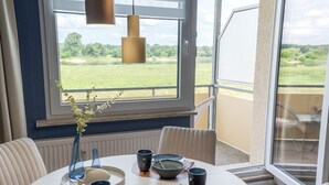 A dining area with a white table set for four, surrounded by white upholstered chairs. A large window with blinds allows natural light and offers a view of a green landscape. - Barby apartment with Elbe view & balcony on the Elbe-Saale Cycle Path (Barby (Elbe))