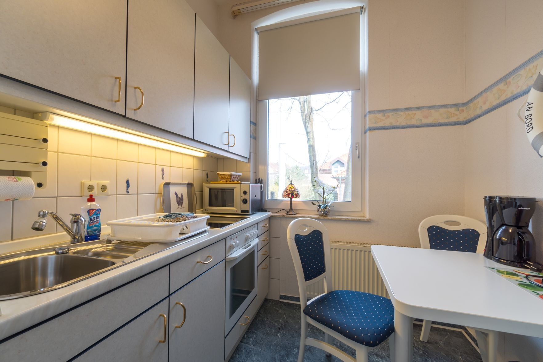 A compact kitchen with a sink, microwave, and stove. There is a window with a view of the outside, a chair, and a table with a coffee maker.