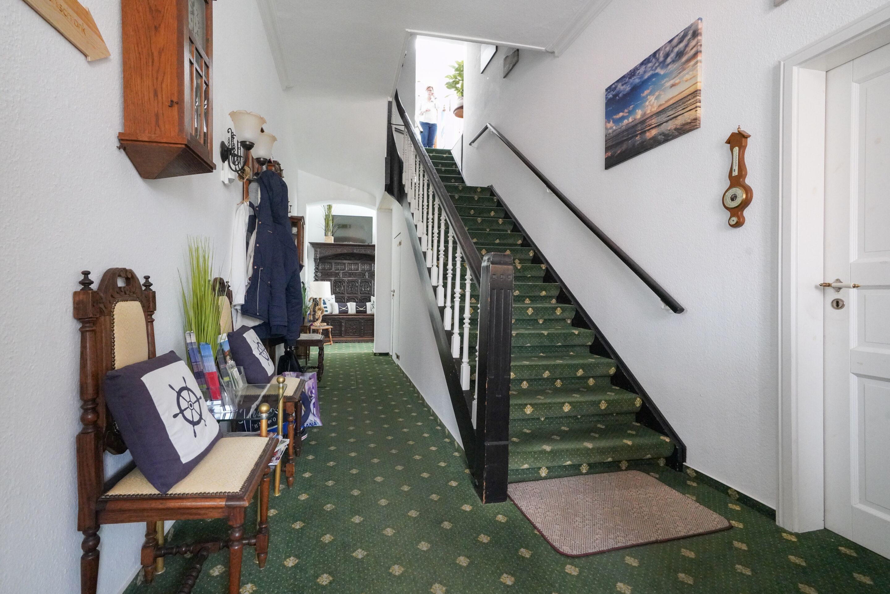 A hallway with a staircase, a wooden chair with a cushion, a wall clock, and a painting.