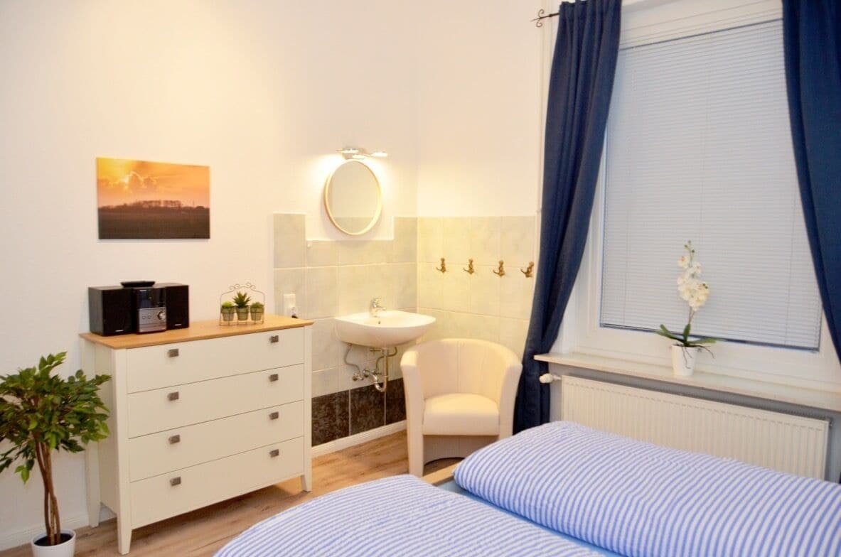 A bedroom with a bed, a bedside table, a dresser, a mirror, a window with curtains, a potted plant, and a wall-mounted radio.