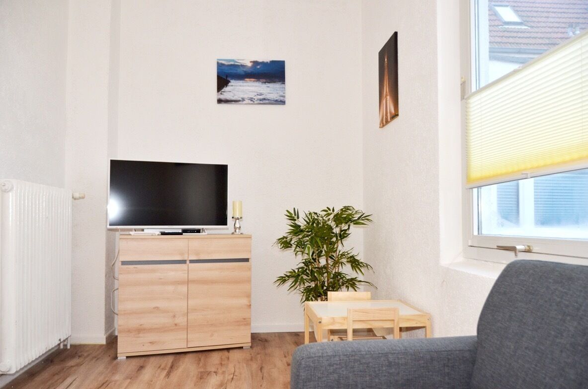 Un salon avec un téléviseur à écran plat posé sur un meuble en bois, une petite table en bois, une plante en pot et un canapé gris.