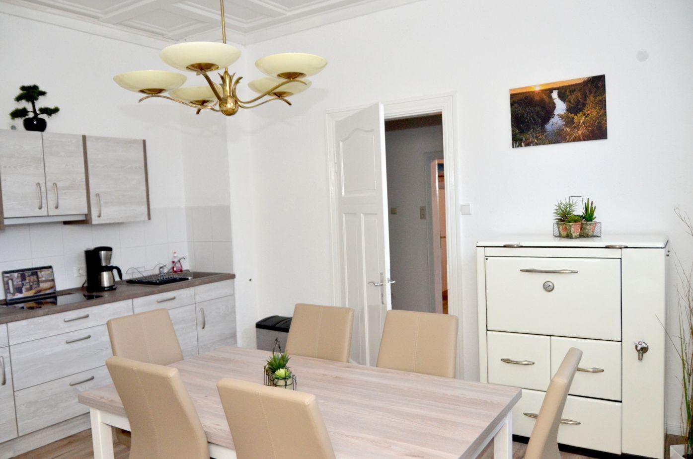 Une cuisine moderne avec des meubles blancs, une table à manger en bois, des chaises beiges, un lustre et un petit meuble orné de plantes.