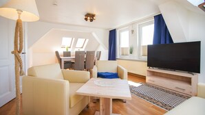 A modern living room with a flat-screen TV, a dining area with chairs, a table, and a large window with curtains.