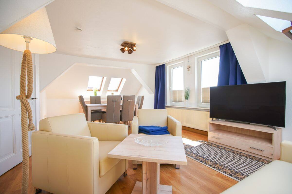 A modern living room with a flat-screen TV, a dining area with chairs, a table, and a large window with curtains.