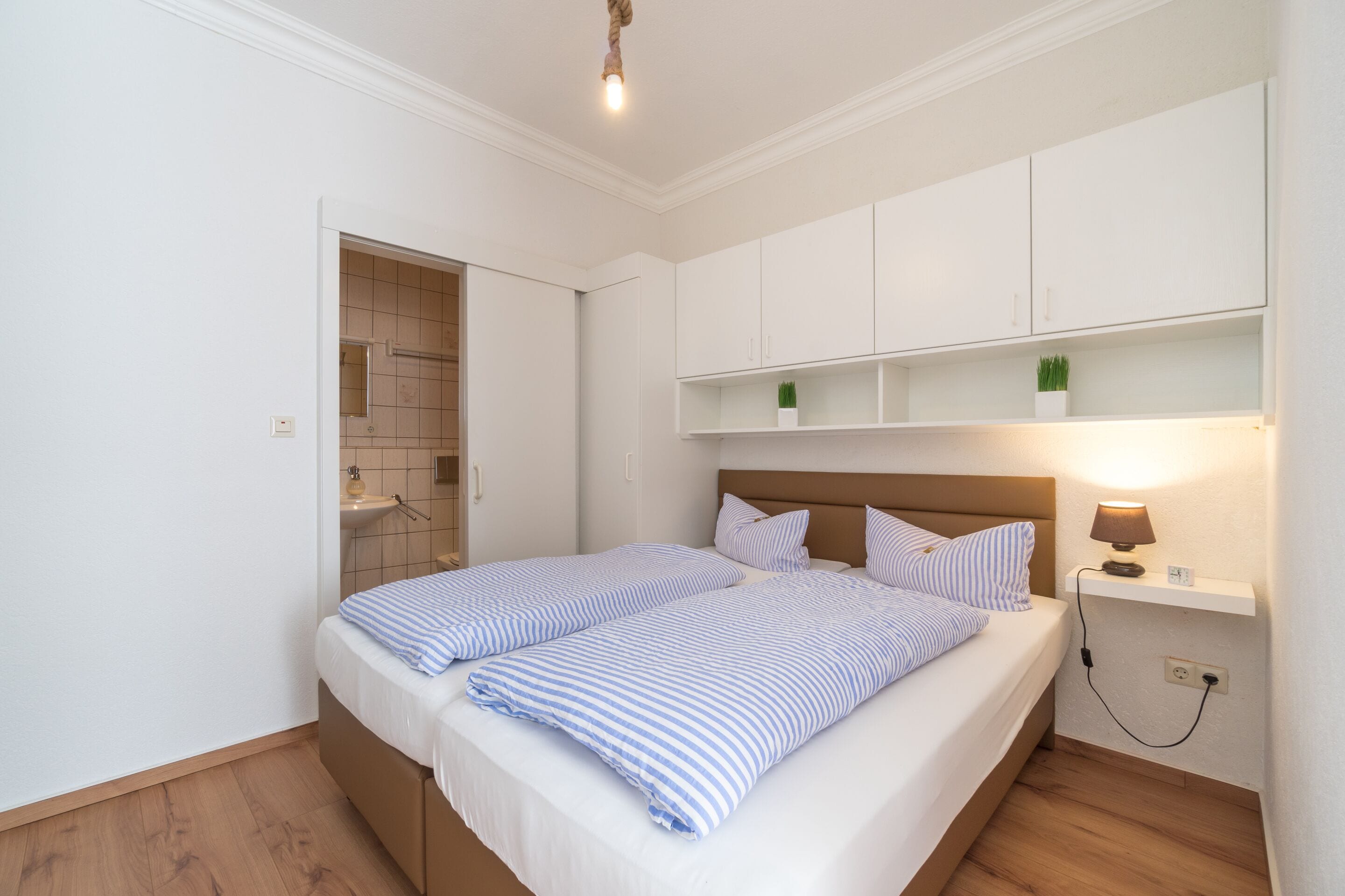 Ein Schlafzimmer mit Bett, Nachttischen, Ablagefläche und einem Regal an der Wand mit Pflanzen sowie einer Badezimmertür.