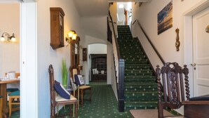A hallway with a green carpet, wooden furniture, a staircase, and a chandelier.