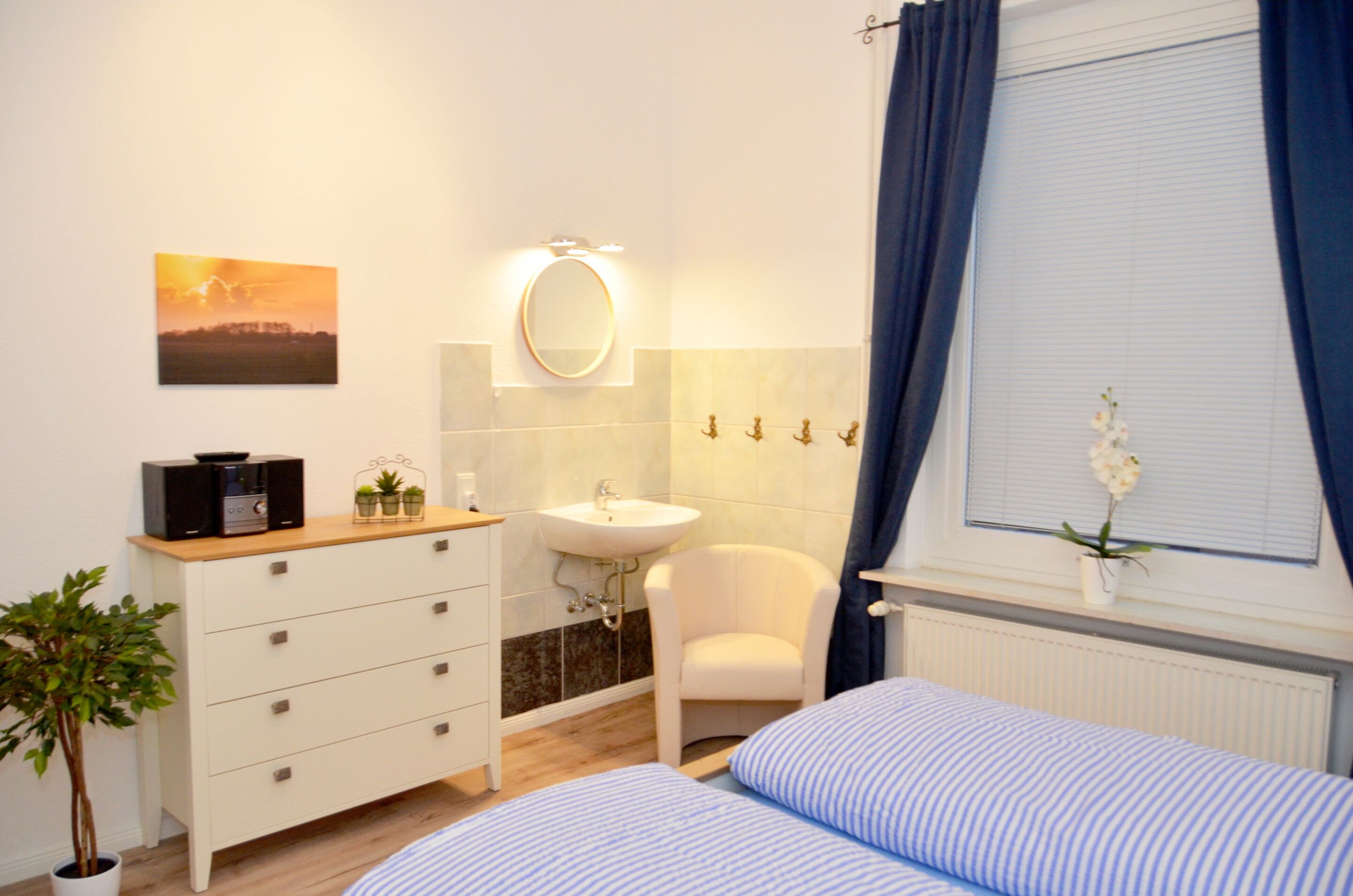 A bedroom with a bed, a bedside table, a dresser, a mirror, a sink, and a window with curtains.