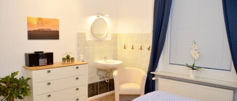 A bedroom with a bed, a bedside table, a dresser, a mirror, a sink, and a window with curtains.