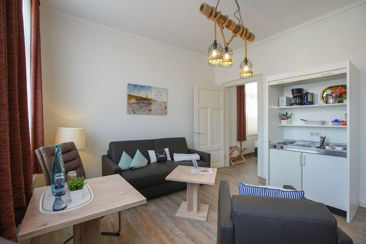 A modern living room with a sofa, a coffee table, and a dining table. There is a kitchen area with a stove and cabinets.