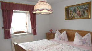 Ein Schlafzimmer mit einem hölzernen Bett, blumigem Bettzeug, einem Nachttisch mit Lampe, einem Fenster mit Vorhängen und einem Bild an der Wand.