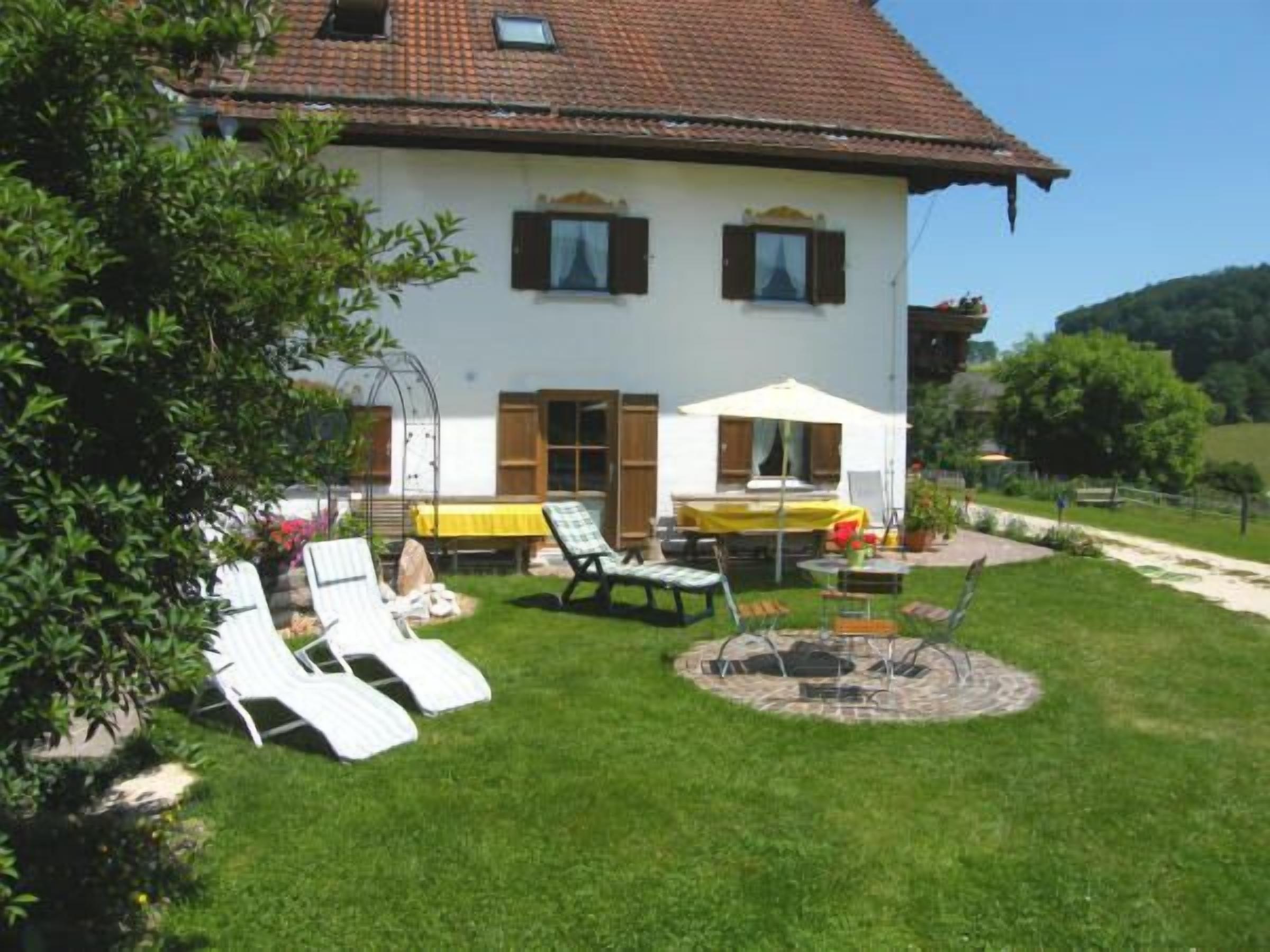 Une maison à deux étages avec un toit en tuiles, un patio couvert avec une table et des chaises, et une pelouse agrémentée de chaises longues.