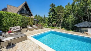 Una piscina amb gandules i una zona d'estar coberta, envoltada d'un jardí ben cuidat i una casa amb teulada inclinada.