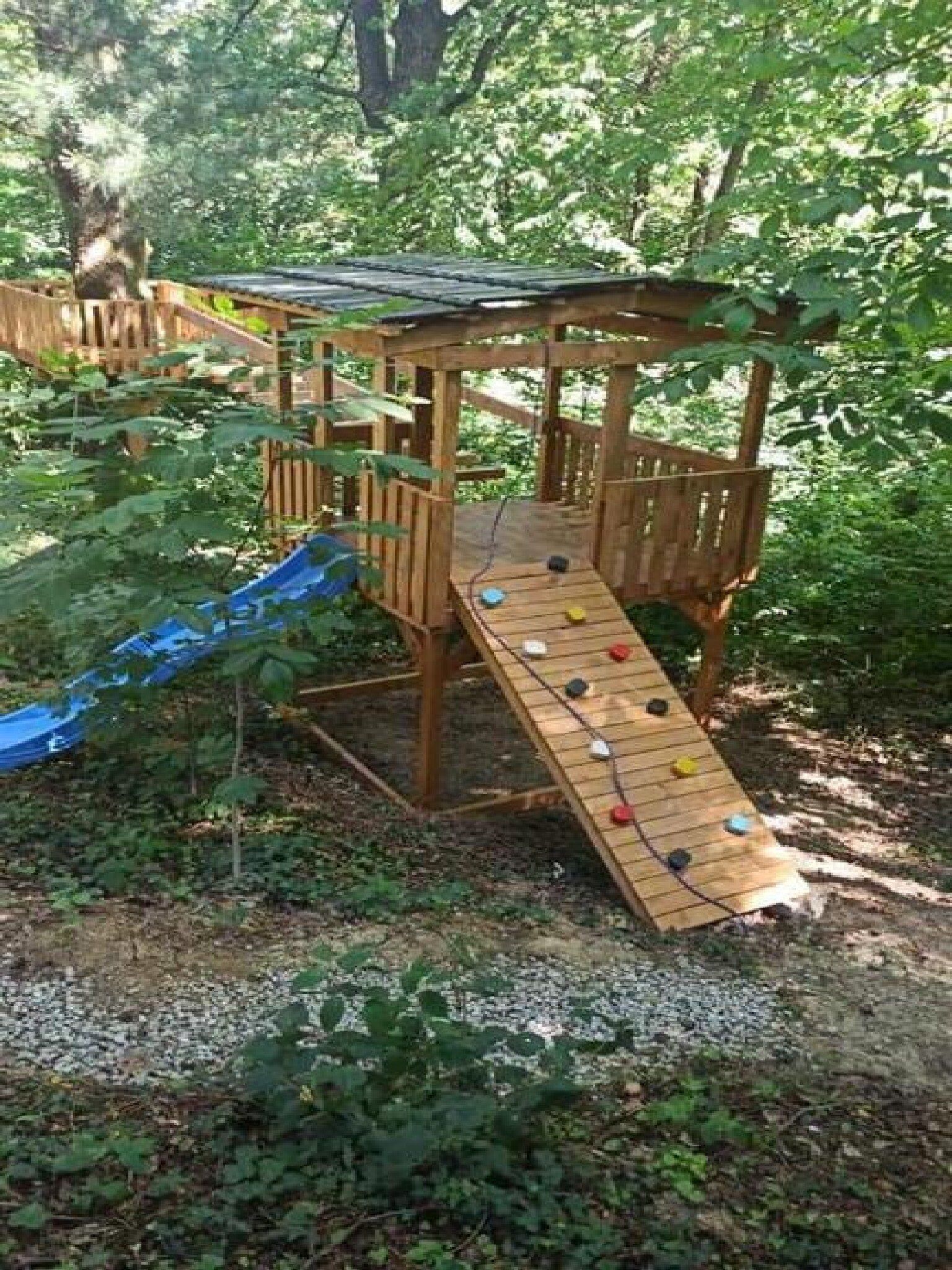 Eine Holzspielanlage mit Rutsche und Kletterwand in einem bewaldeten Gebiet.