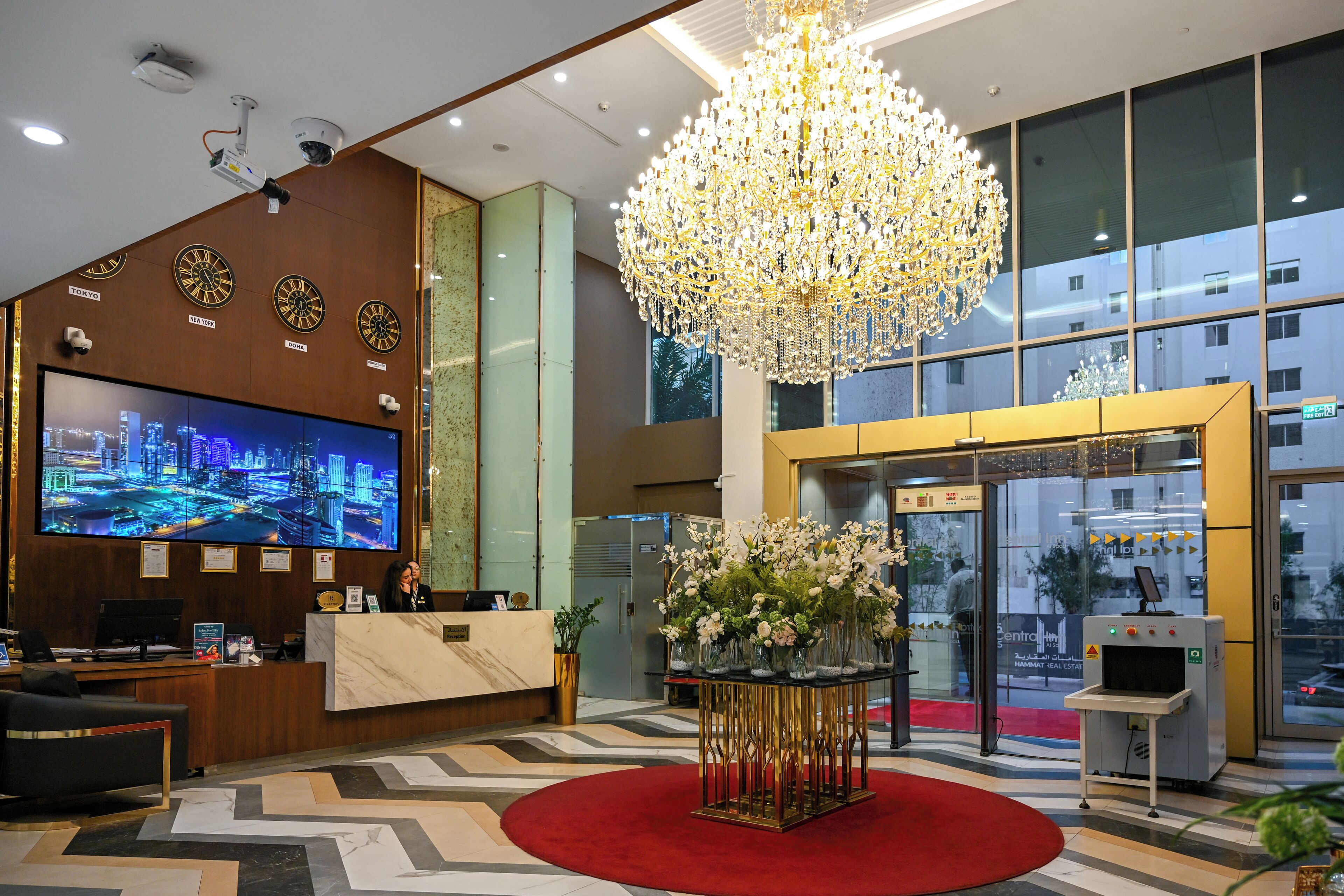 Ein Hotel-Empfangsbereich mit einem großen digitalen Bildschirm, der eine Stadtansicht zeigt, einer prunkvollen Kristallleuchtre und einem Empfängertresen mit rotem Teppich.