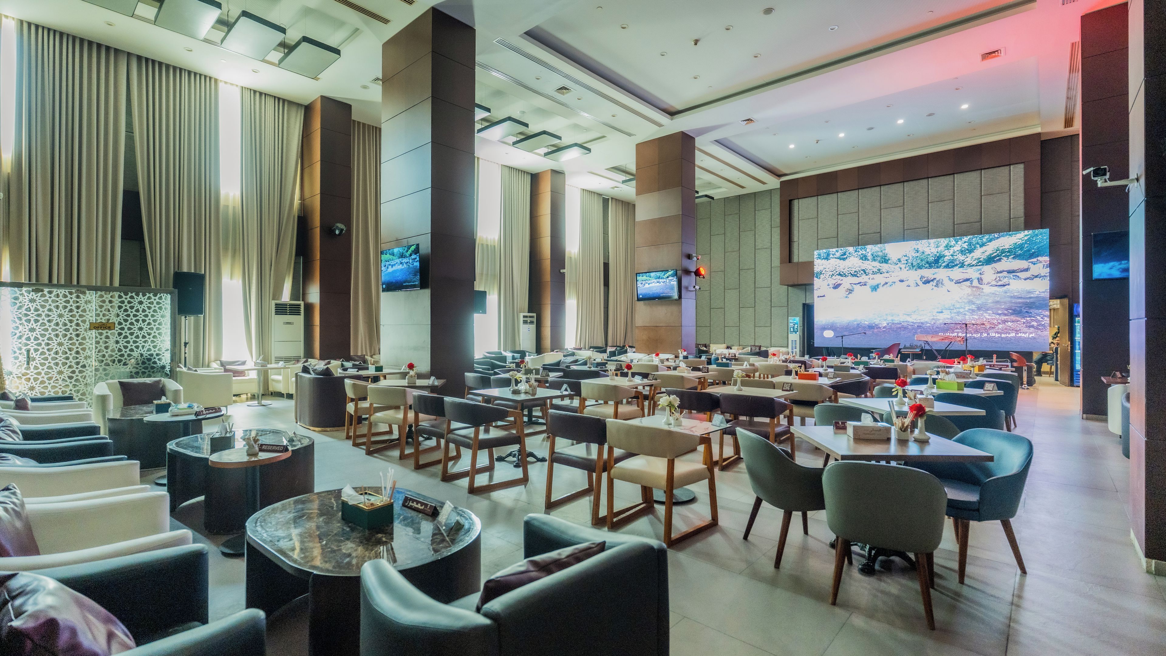 Een ruime hotellobby met verschillende zithoeken, grote schermen en een modern design.