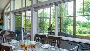 A dining area with a large table set for four, surrounded by large windows offering a view of a lush garden.