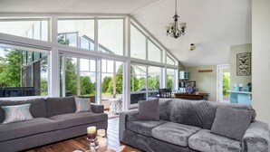 A spacious living room with large windows, a grey sofa set, a wooden coffee table with candles, and a chandelier.