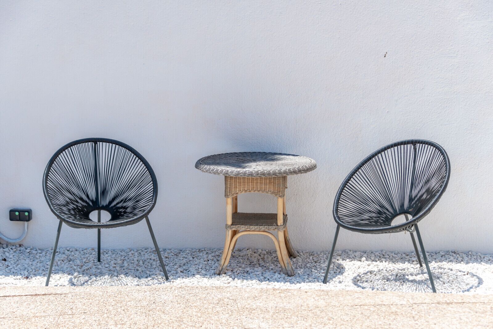 Three black wireframe chairs and a wicker table against a white wall.