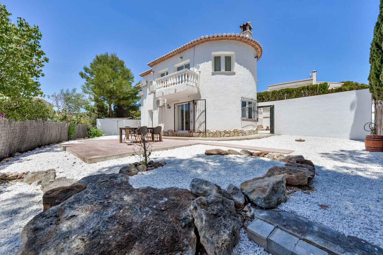 A two-story house with a white exterior and a tiled roof. There is a patio with a table and chairs, and a small garden area with rocks and a potted plant.