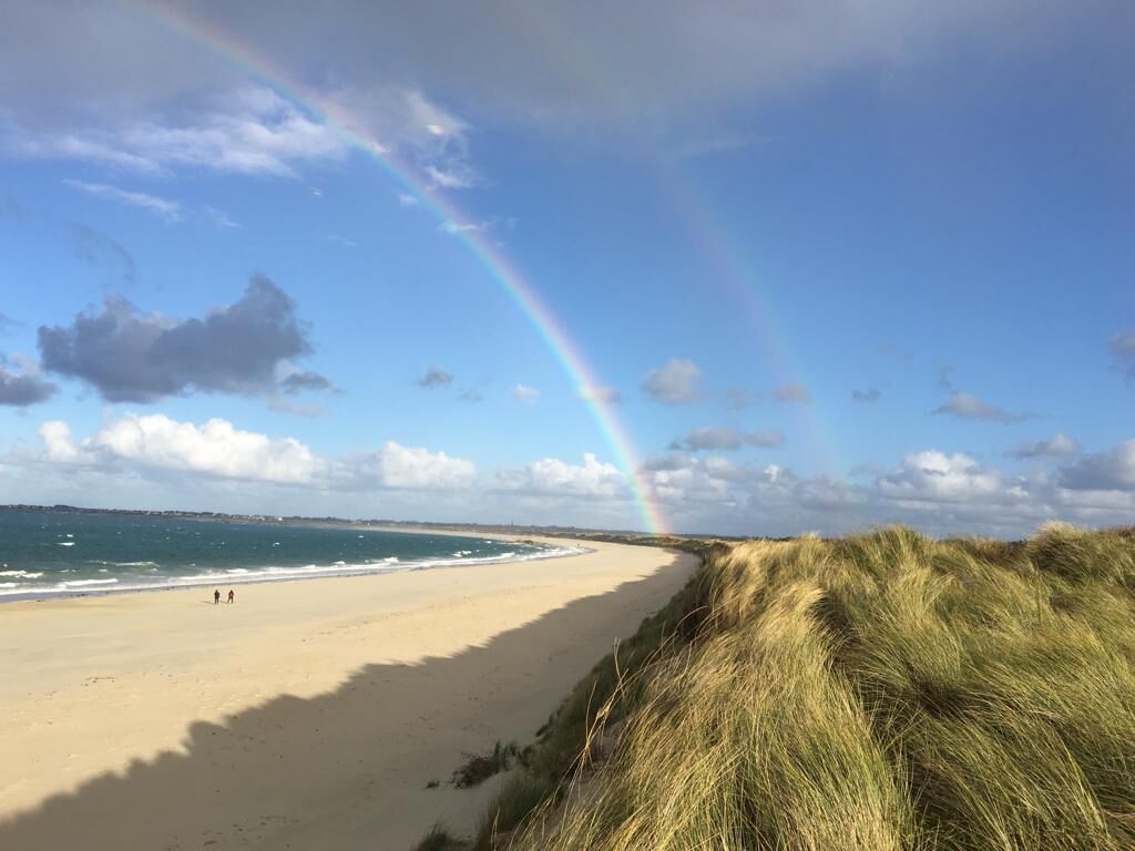 Una playa con un arcoíris visible, orilla arenosa y dunas cubiertas de hierba.
