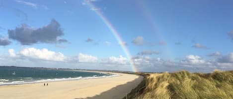 Una playa con un arcoíris visible, orilla arenosa y dunas cubiertas de hierba.