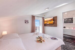A hotel room with a bed, a bedside table with a lamp, a window with curtains, a painting on the wall, and a bathroom with a mirror and toiletries. - Pool Apartments Crikvenica (Crikvenica)