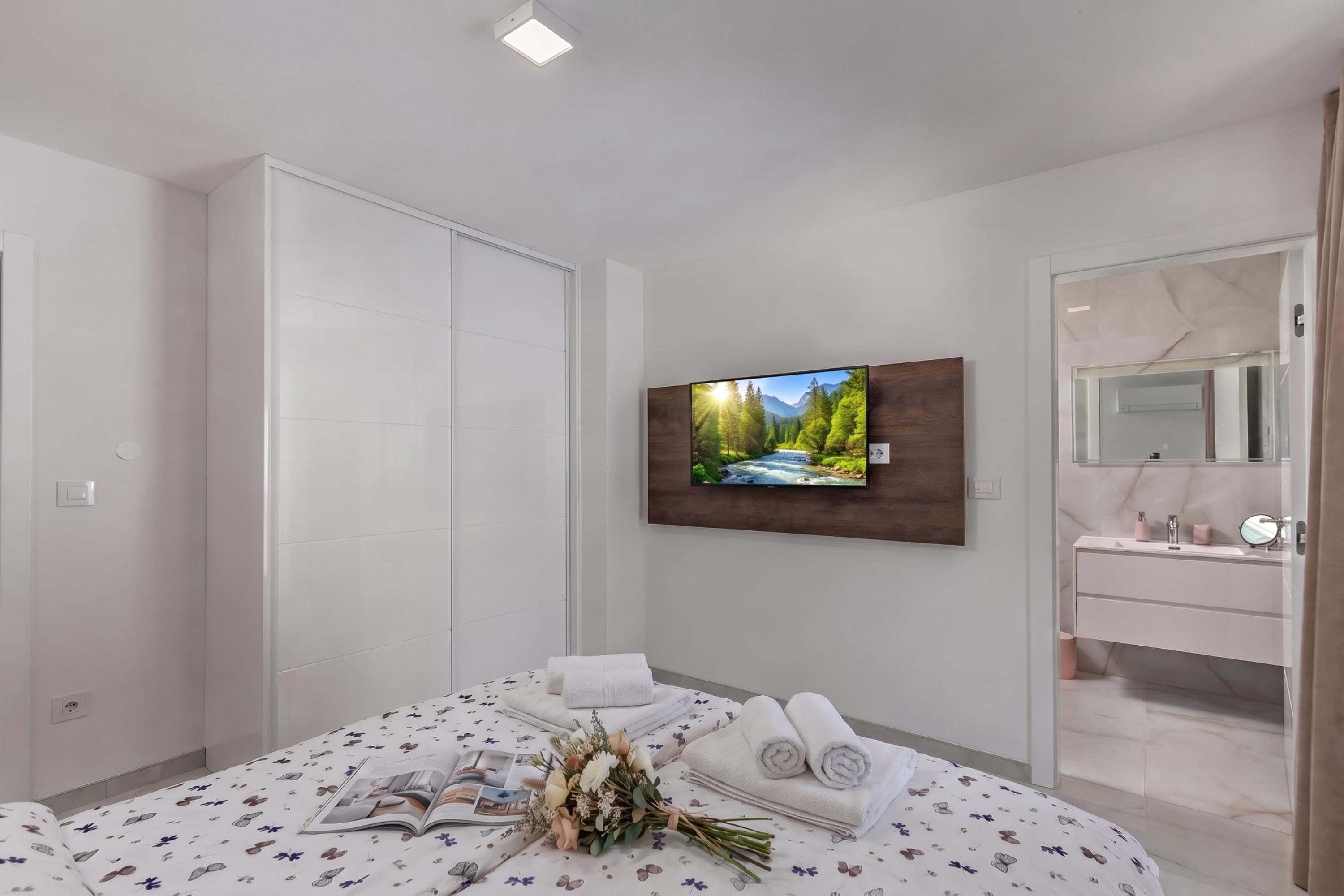 A modern bedroom with a bed, a flat-screen TV, a vanity with a mirror, and a view of a mountain landscape.