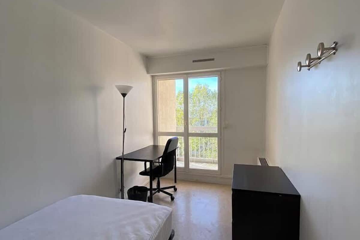 A small, bright room with a bed, a desk, and a chair.