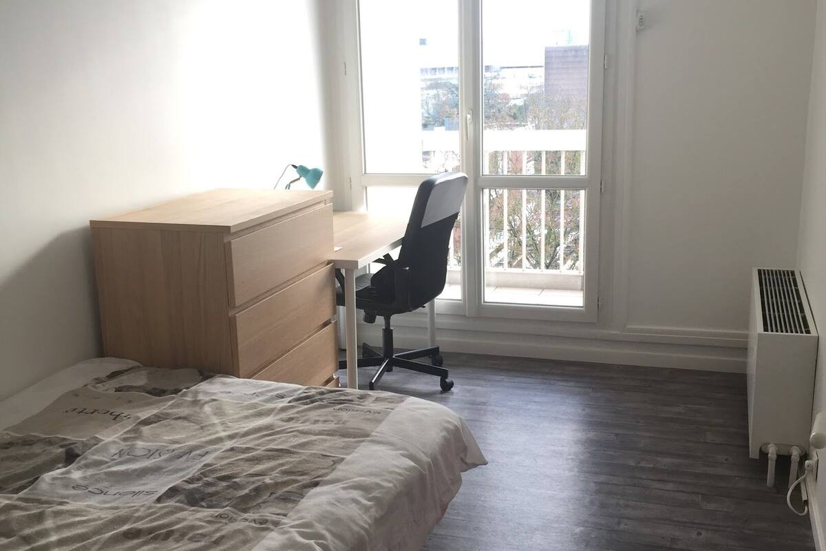 A bedroom with a bed, a wooden dresser, a desk with a chair, a radiator, and a balcony with a view of buildings.
