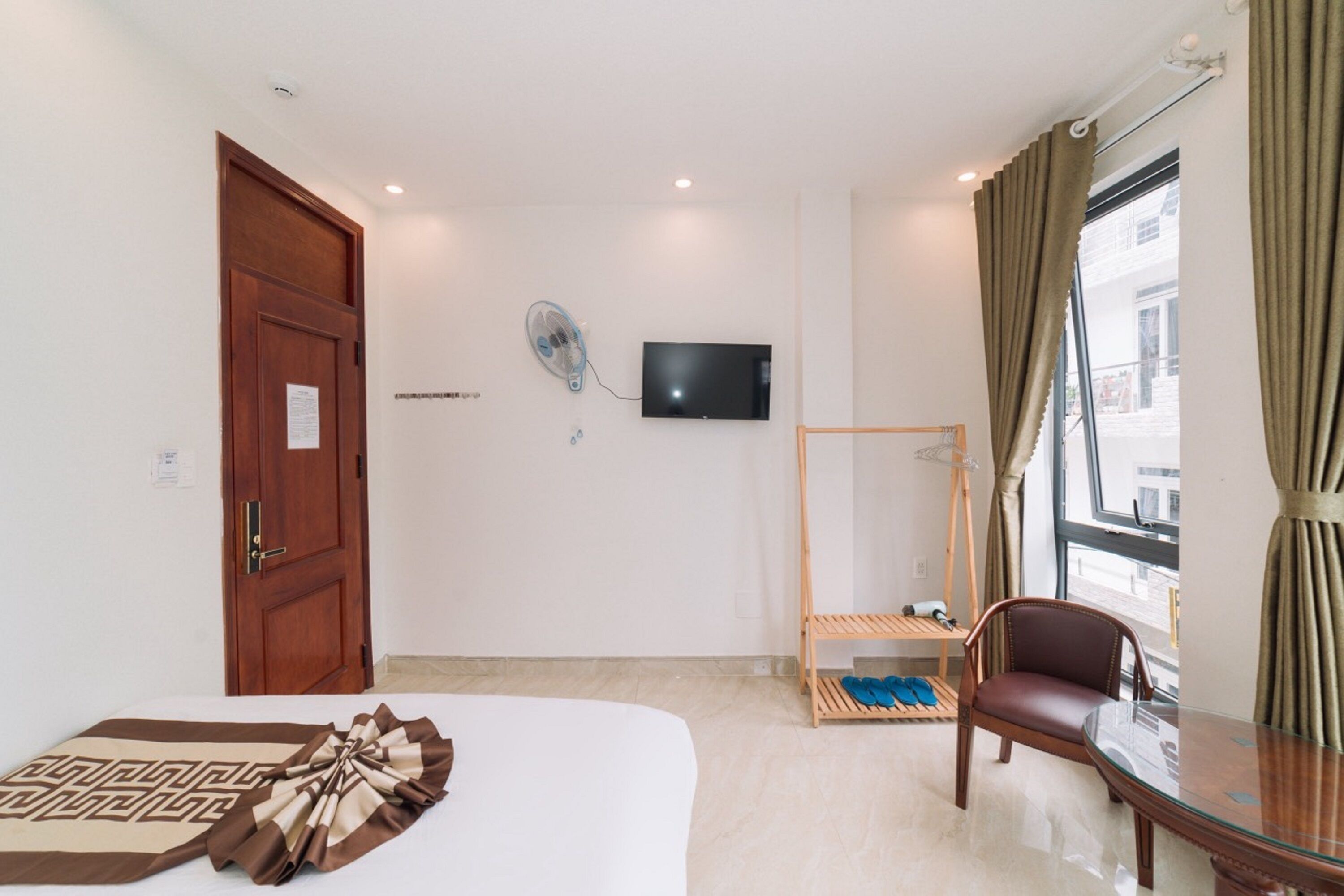 A hotel room with a bed, a TV mounted on the wall, a wooden chair, a small table, and a window with curtains.