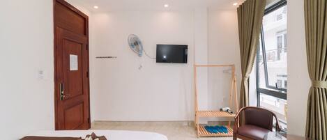 A hotel room with a bed, a TV mounted on the wall, a wooden chair, a small table, and a window with curtains.