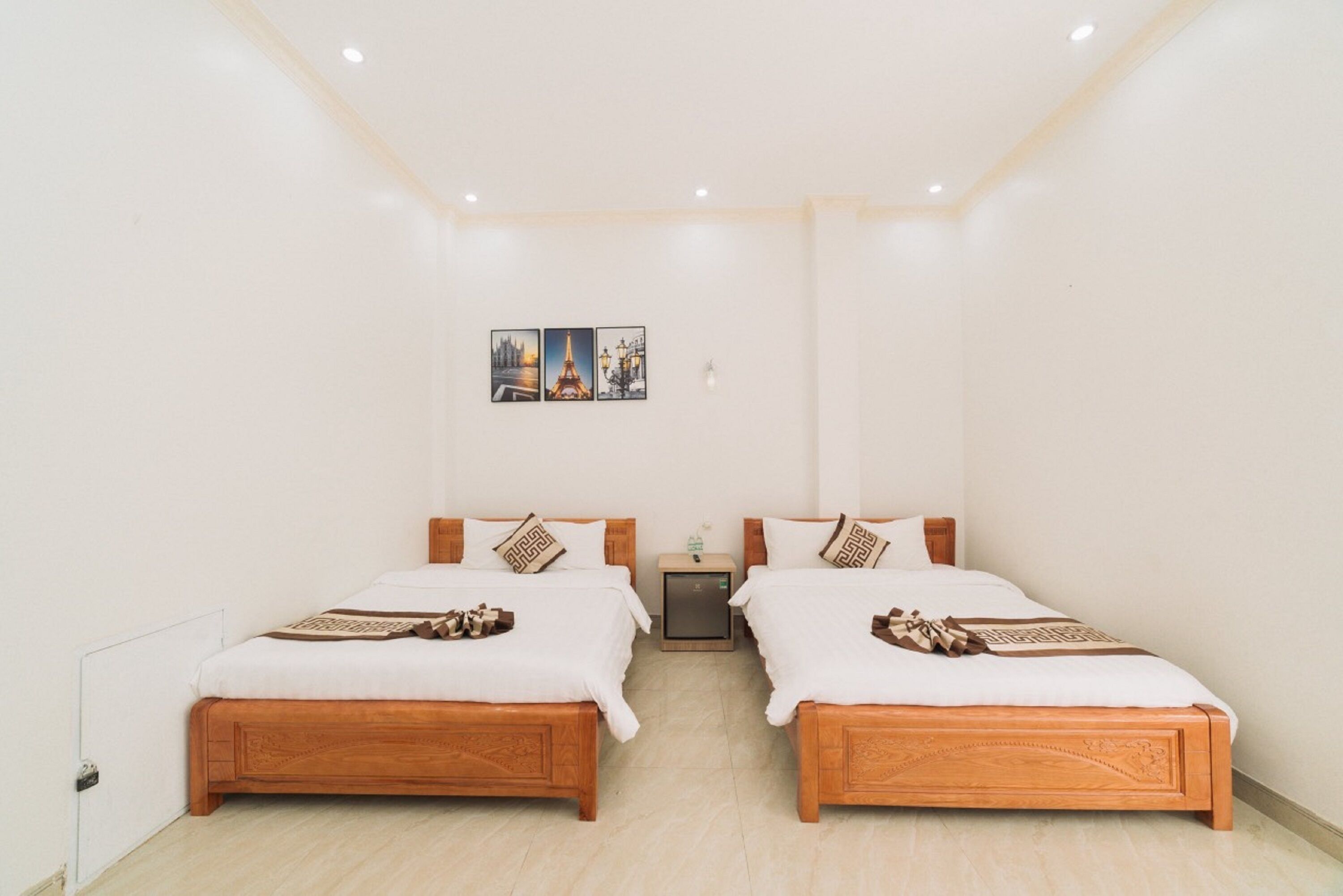 A hotel room with two wooden beds, a small bedside table, a nightstand, and a wall-mounted artwork depicting a cityscape.