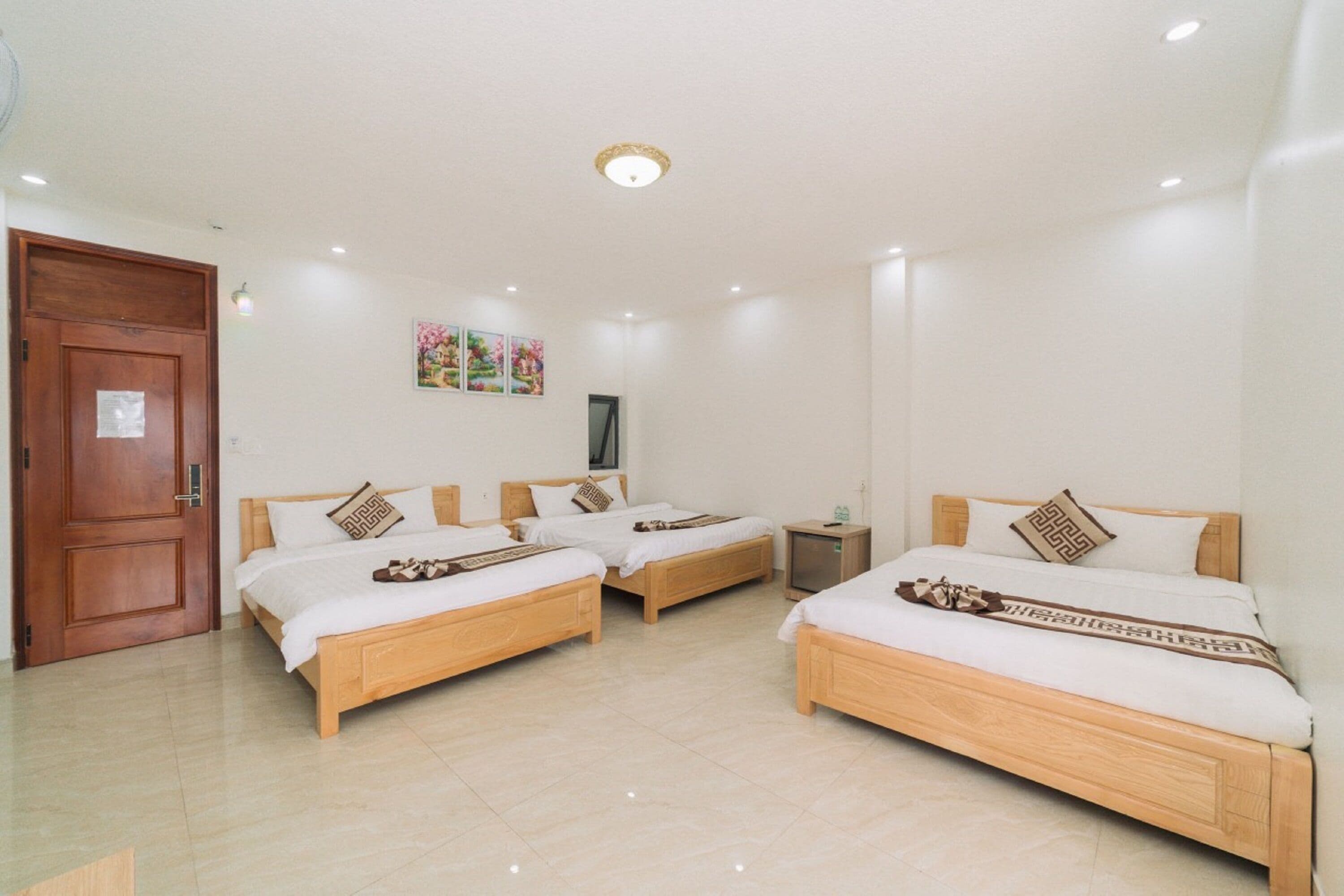 A hotel room with three wooden beds, a wooden door, a small bedside table, and a wall-mounted TV.
