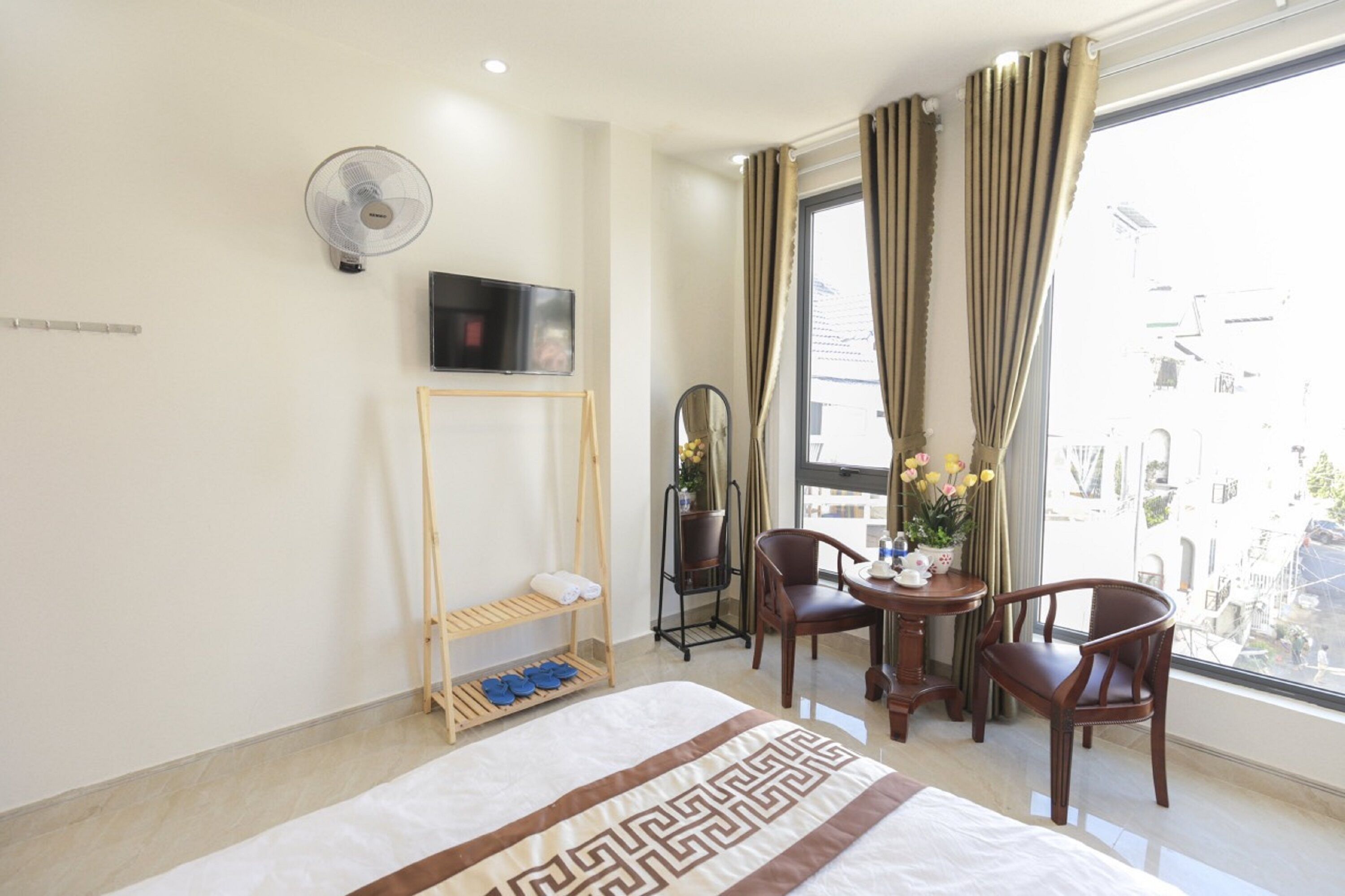 A hotel room with a bed, a TV, a fan, a small table with chairs, a window with curtains, and a view of the city.