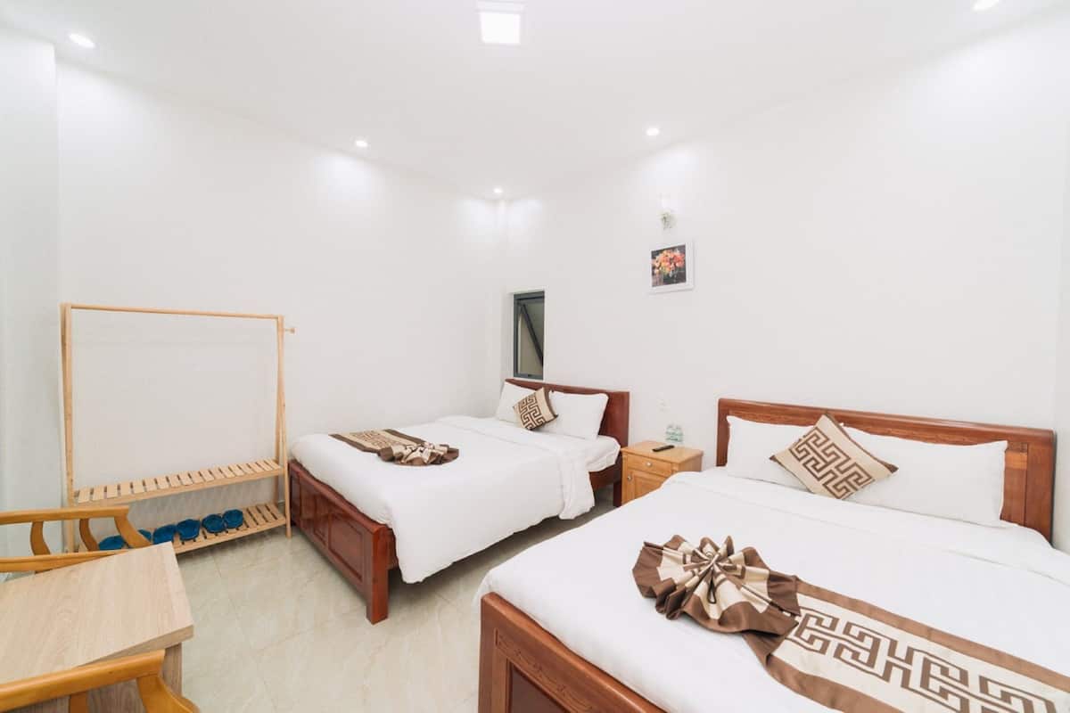 Quarto de hotel com duas camas, uma cadeira de madeira, sapateira e uma pequena mesa.