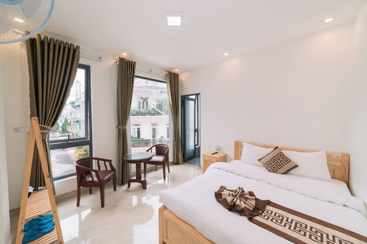 Quarto de hotel com uma cama, uma cadeira de madeira, uma mesa pequena e uma janela com cortinas.