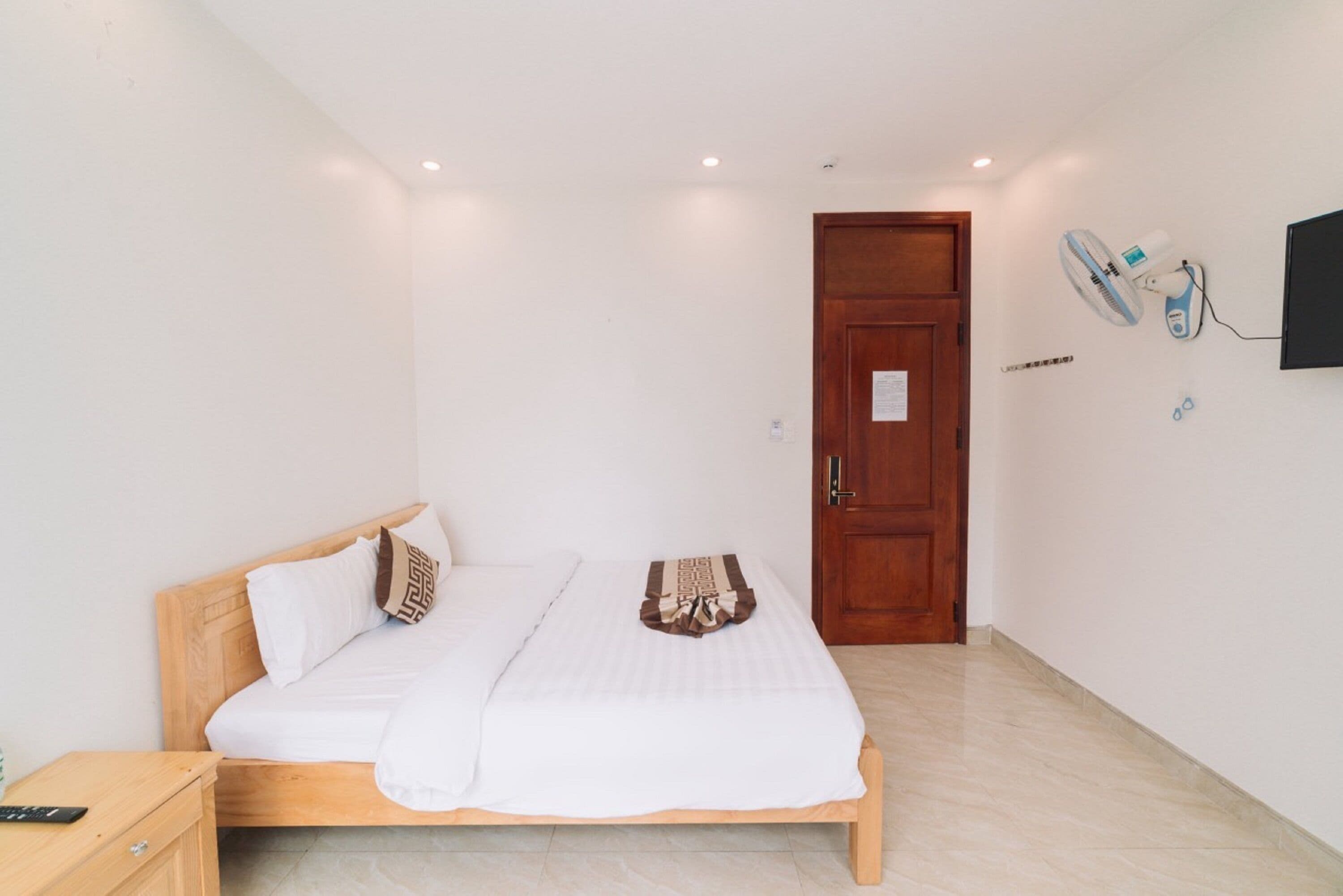 A bedroom with a wooden bed, white bedding, a wooden nightstand, a wall-mounted fan, and a door.