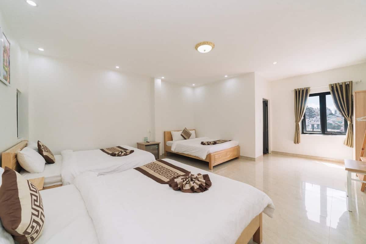Quarto de hotel com três camas, cabeceira de madeira, mesa de cabeceira pequena e janela com cortinas.