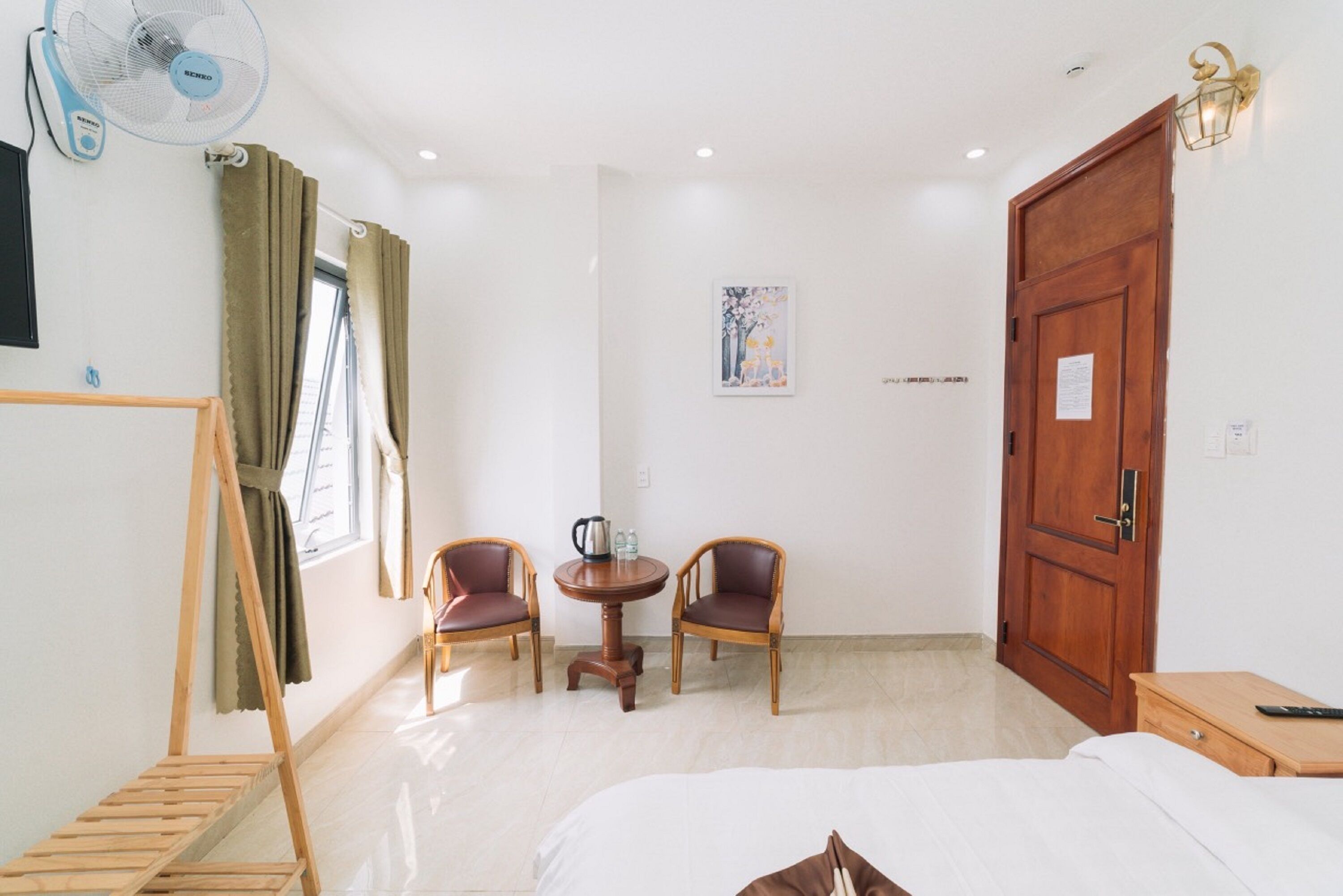 A hotel room with a bed, a wooden side table, two chairs around a small table, a standing fan, a wardrobe, and a painting on the wall.