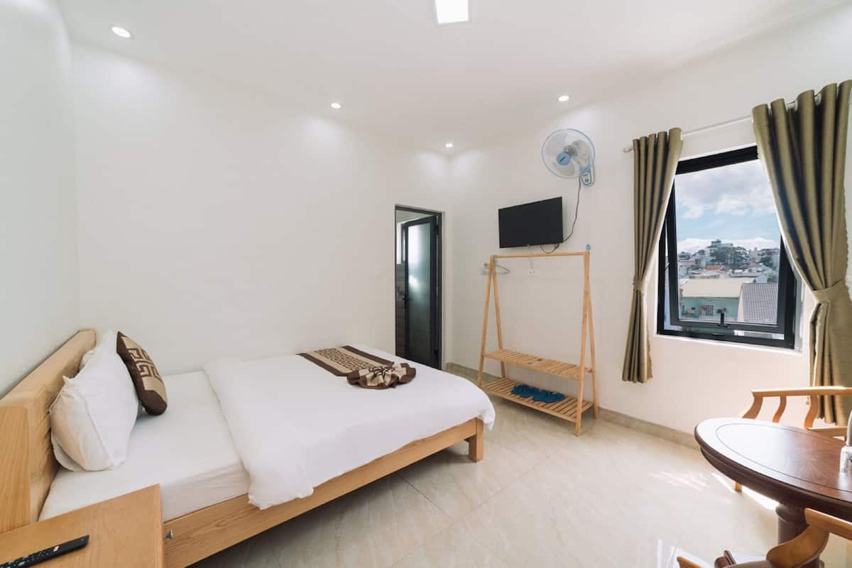 Quarto de hotel com cama, televisão fixada na parede, mesa pequena com cadeiras e janela com cortinas.