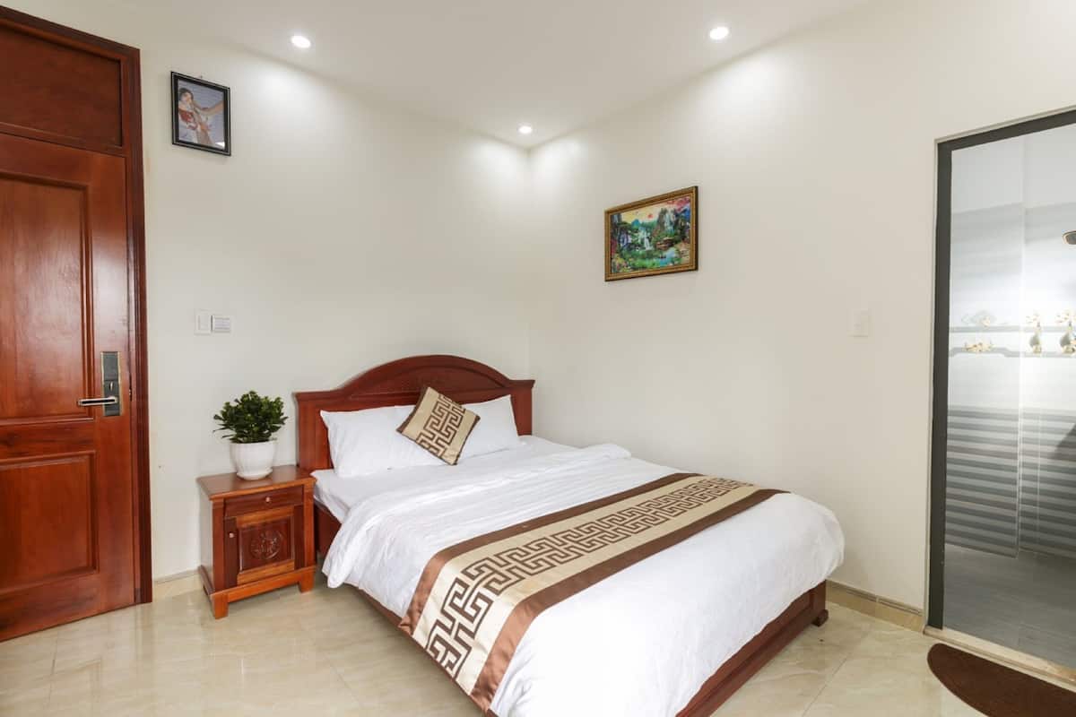 Cama bem arrumada com colcha estampada, mesa de cabeceira de madeira com uma planta e uma porta à esquerda.