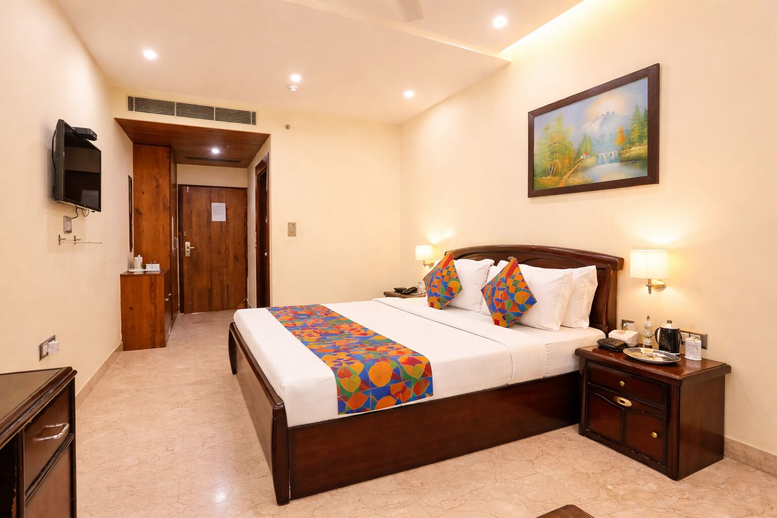 A hotel room with a bed, bedside tables, a TV, a painting, and a door.