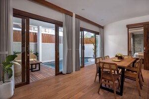A spacious room with a wooden floor, a dining table with chairs, a large sliding glass door leading to a pool area, and a potted plant.